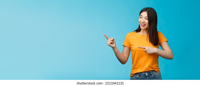 Friendly enthusiastic asian girl curiously looking sideways, pointing left interesting object, grinning joyfully discuss new opened store, stand blue background carefree lively talking - Shutterstock ID 2211041131