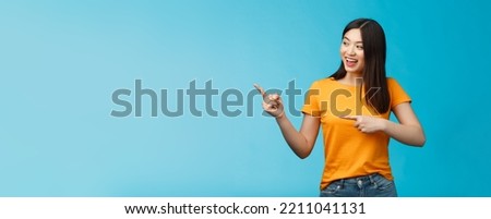 Friendly enthusiastic asian girl curiously looking sideways, pointing left interesting object, grinning joyfully discuss new opened store, stand blue background carefree lively talking