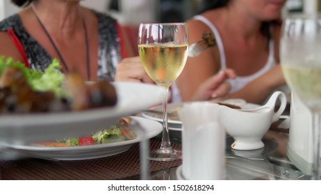 Friendly Or Family Dinner. In The Spotlight, Someone Pours Wine Into A Glass From A Bottle. People Eat And Drink At A Big Table