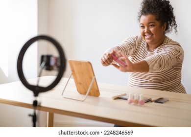 Friendly female beauty blogger curly hispanic overweight young lady sitting at vanity table at home, showing pink lip gloss at smartphone camera, influencer making review, recommend beauty product - Powered by Shutterstock