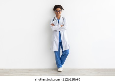 Friendly Female Black Doctor In Glasses Posing With Folded Arms Looking At Camera Standing Leaning On Wall, Lady Wearing Blue Uniform, Coat And Stethoscope, Full Body Length, White Studio Background