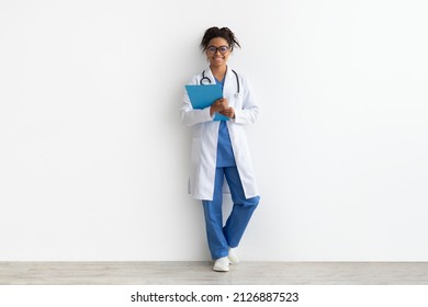 Friendly Female Black Doctor In Glasses Holding Clipboard Looking At Camera Standing Leaning On Wall, Lady Wearing Blue Uniform, Coat And Stethoscope, Full Body Length, White Studio Background