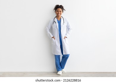 Friendly Female Black Doctor Posing With Hands In Pockets Looking At Camera Standing Leaning On Wall, Lady Wearing Blue Uniform, Coat And Stethoscope, Full Body Length, White Studio Background Banner