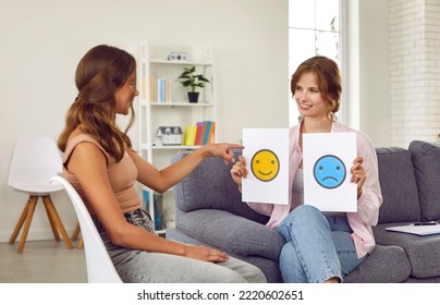 Friendly female child psychologist talks to teenage girl about emotions she is currently feeling. Psychologist shows teenage girl two sheets of paper with positive and sad emoticons drawn on them. - Shutterstock ID 2220602651