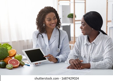 Friendly female dietologist showing black lady patient empty digital tablet screen in her office, technologies in medical care - Shutterstock ID 1687429024