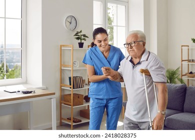 Friendly female nurse helping an injured senior man walk with a crutch, holding his hand, supporting him. Treatment and rehabilitation after injury, assisted living facility, senior care concept - Powered by Shutterstock