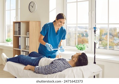 Friendly female nurse taking care of man in hospital. Happy young woman in scrubs holding clipboard, standing by bed with vitamin therapy IV drip, talking to male patient and smiling  - Powered by Shutterstock