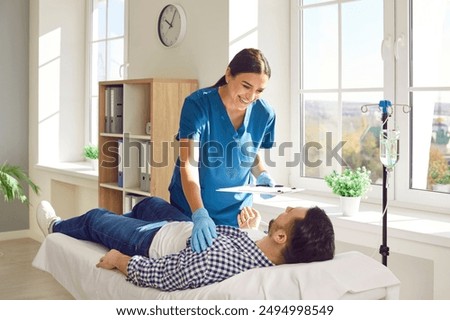 Friendly female nurse taking care of male patient at clinic. Smiling young woman in scrubs holding clipboard and standing by bed with man receiving vitamin therapy IV drip