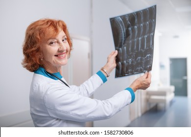 Friendly Female Oncologist Smiling To The Camera, Holding MRI Scan Of A Patient. Elderly Practitioner Examining MRI Scan Of A Patient