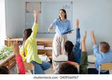 Friendly female teacher in classroom teaches elementary school students, kids raising hands to teacher aasks questions to small students sitting at their desks - Powered by Shutterstock