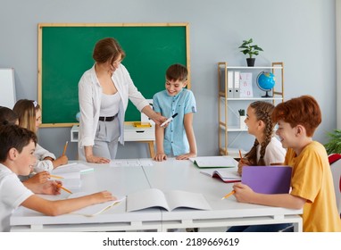 Friendly Female Teacher Conducts Creative Lesson For Students Of Modern Private School. Teacher Gives Marker To Student In Front Of Children Sitting In Circle At Large Desk With Notebooks And