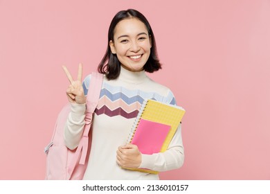 Friendly fun teen student girl of Asian ethnicity wear sweater backpack hold books showing victory sign isolated on pastel plain light pink color background. Education in university college concept