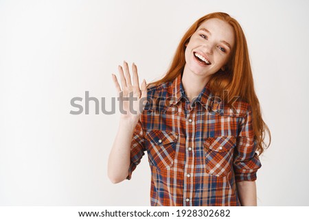 Friendly girl with ginger hair and blue eyes greeting, saying hi and waving hand, standing over white background.