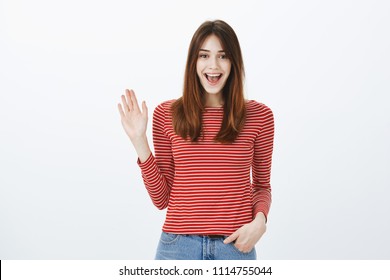 Friendly Girl Saying Hi To Best Friends. Portrait Of Cheerful Bright European Woman In Striped Pullover, Waving Hand In Hello Gesture And Smiling Broadly, Pleased To See Familiar Person On Street