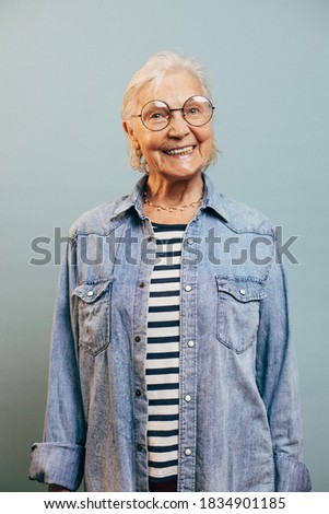 Friendly good looking senior woman in casual clothes denim jacket and striped shirt. Round glasses. White sort hair. Smart style concept. Isolated over blue background