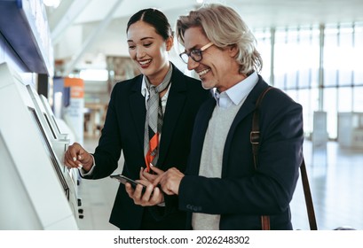 Friendly Ground Attendant Helping Male Traveler In Doing The Self Check-in Using A Machine At The Airport Terminal. Businessman At Self Service Check In Machine With Airport Female Staff.