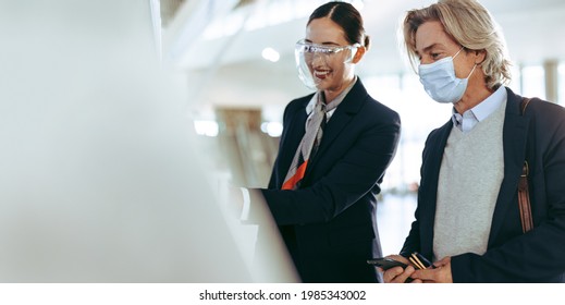 Friendly Ground Attendant Wearing Face Shield Helping Male Travel With Face Mask In Doing The Self Check-in Using A Machine At The Airport Terminal. Safe Air Travel Post Pandemic Lockdown Reopening.