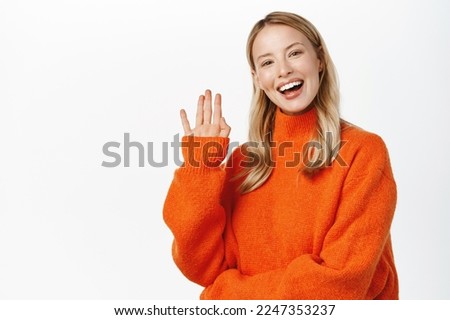 Friendly happy blond girl say hello, waving hand and smiling, greeting, welcome gesture, standing in red sweater against white background