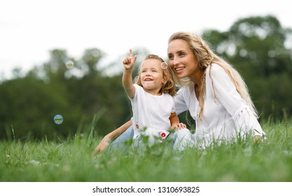 Friendly And Happy Family In Nature.Positive And Beautiful Young Mother And Her Little Mischievous Daughter Laughing Soap Bubbles Sitting On The Grass For A Walk In The Park.Summer.Childhood.Lifestyle