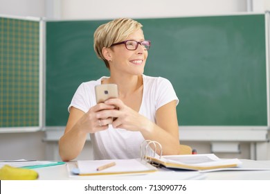 Friendly Happy Young University Student Sitting Working At A Table In The Classroom Holding Her Mobile And Looking To The Side With A Smile