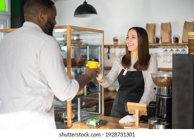 Friendly and helpful waitress is serving an African-American male customer, preparing and giving paper cup of coffee to multiracial guy. Woman in apron holds out take-away coffee to guest of cafe - Powered by Shutterstock