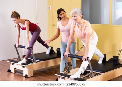 Friendly Hispanic Woman Professional Pilates Instructor Assisting Elderly Female To Do Exercises On Reformer. Active Lifestyle Concept