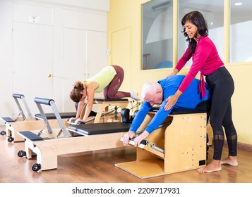 Friendly Hispanic Woman Professional Pilates Instructor Helping Senior Man Exercising On Combo Chair In Fitness Studio