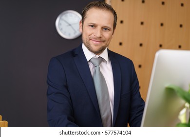 Friendly Hotel Manager With A Warm Welcoming Smile Standing Behind His Computer At The Front Desk Of A Luxury Hotel