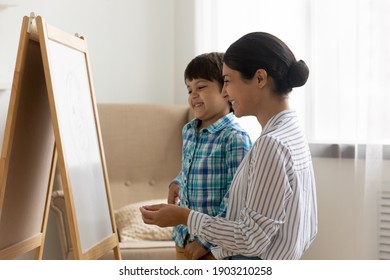 Friendly indian woman psychologist   small boy stand by easel board hold crayon planning picture talk visual art therapy session  Happy hindu mother teach little son to draw using piece chalk