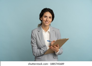 Friendly instructor or agent woman wear grey blazer and white blouse hold folder, pen looking at camera. Smiling female office worker isolated on studio blue background. 