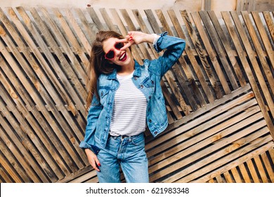 Friendly laughing girl in jeans suit making a peace sign. Smiling female model in striped sweatshirt posing next to wooden fence background. Charming lady with new denim outfit. Happy young woman - Powered by Shutterstock