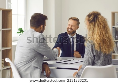 Friendly lawyer, realtor or financial advisor shakes hands with his clients to young couple. Husband and wife who want to buy house apply to real estate agency, insurance broker or bank employee.