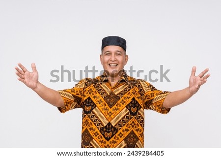 Friendly local Indonesian man wearing a traditional batik shirt and kopiah hat, offering a warm welcome with open arms. Isolated on white.