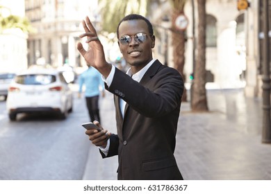 Friendly looking successful young Afro American entrepreneur in elegant black suit and eyewear texting on cell phone and raising hand while hailing up cab, standing on street in urban surroundings - Powered by Shutterstock