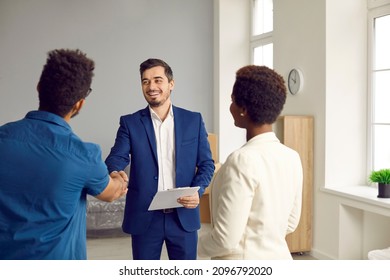 Friendly Male Real Estate Agent Shaking Hands And Welcoming Young Family In New Home. African American Couple Standing With Their Backs To Camera While Talking To Realtor. Housing Rental Concept.
