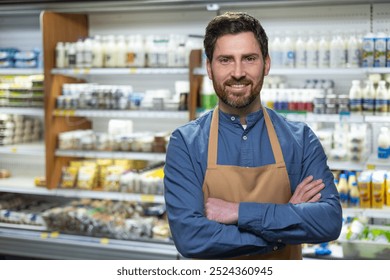 Friendly male supermarket employee with apron standing confidently in front of dairy section. Shelves filled with various products in background, showcasing retail environment. - Powered by Shutterstock