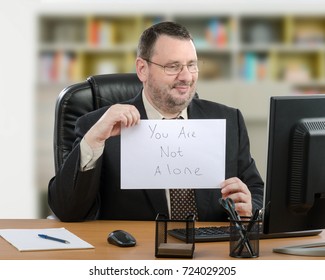 Friendly Male Telemedicine Psychiatrist Shows Written Message You Are Not Alone To Desktop Monitor During Video Consultation Online. 55 Years Old Bearded Man Wearing Black Suit Sits At Wooden Desk.