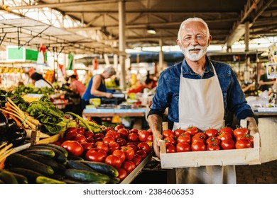 Friendly man tending an organic vegetable stall at a farmer's market and selling fresh vegetables. Male gardener selling basket full of fresh tomatoes at the vegetable market. - Powered by Shutterstock