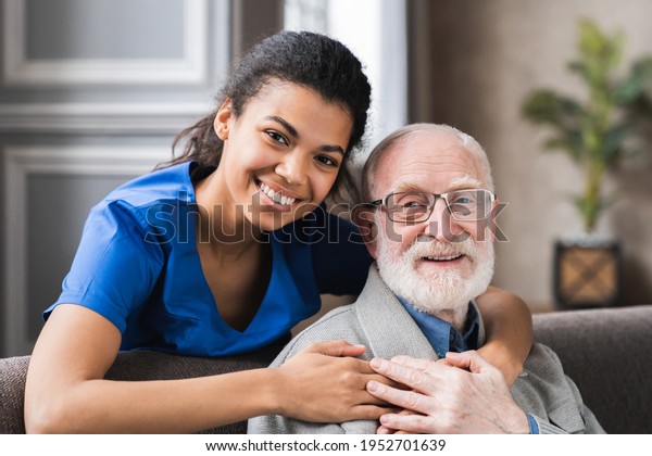 Friendly mature general practitioner communicating
with pleasant 80s male patient, sitting together on sofa. Smiling
trustful young doctor giving psychological help to elder man at
home visit.