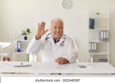 Friendly Mature Male Doctor At Desk Waving Hand Welcoming Clients In Hospital Office. Happy Senior Physician Looking At Camera Saying Hello Or Goodbye To Patients On His Medical Video Blog Or Channel