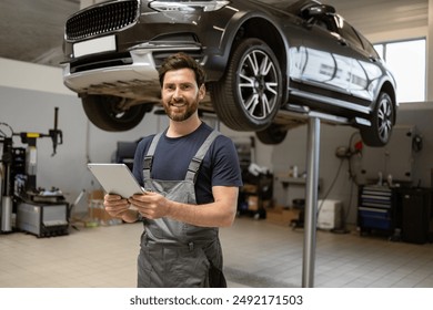 Friendly mechanic using tablet in modern car repair shop with car on lift. Professional technician checking vehicle diagnostics and maintenance. Concept of automotive industry and customer service. - Powered by Shutterstock