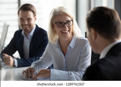 Friendly middle aged female leader laughing at group business meeting, happy old businesswoman enjoying fun conversation with partner, smiling mature business coach executive talking to colleague
