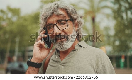Friendly middle aged man with gray hair and beard wearing casual clothes using his mobile phone. Mature gentleman in eyeglasses talking on cell phone outdoors