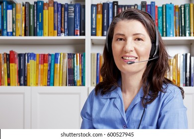 Friendly Middle Aged Woman With Headset And A Blue Shirt In Front Of Lots Of Books, Talking To A Customer Via The Internet, Webcam View, Copy Space