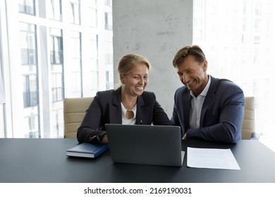 Friendly middle-aged male female workmates sit at desk in modern office looking cheerful work together on corporate task use laptop. Teamwork, workflow, new software, fun at workplace use tech concept