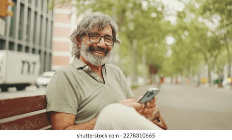 Friendly middle-aged man with gray hair and beard wearing casual clothes sits on bench. Mature gentleman in eyeglasses looks at the camera and smiles - Powered by Shutterstock