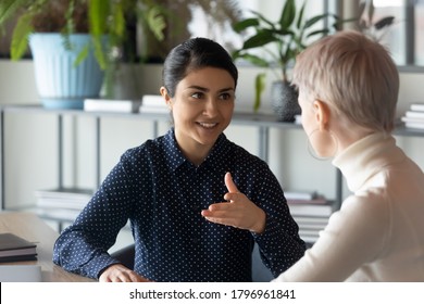 Friendly Millennial Indian Businesswoman Talking To Blonde Female Colleague, Sitting At Table In Office. Two Young Multiracial Employees Discussing Working Issues Or Enjoying Informal Conversation.