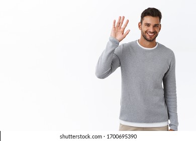 Friendly modest handsome bearded man in grey sweater saying hi with hesitation lifting one palm, waving hand in hello, greeting gesture, smiling cute, standing white background, meet new people