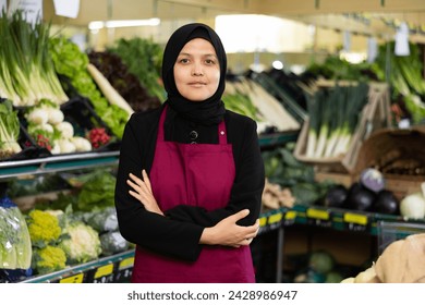 Friendly Muslim woman seller in hijab and apron stands with arms crossed on chest and waits for customers.Vegetable shop next to home,hypermarket, supermarket vegetable department - Powered by Shutterstock