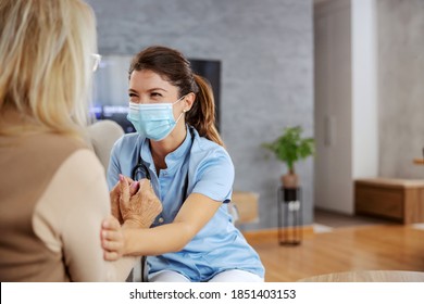 Friendly Nurse With Protective Mask On Face Sitting At Home With Senior Woman And Comforting Her During Corona Virus.